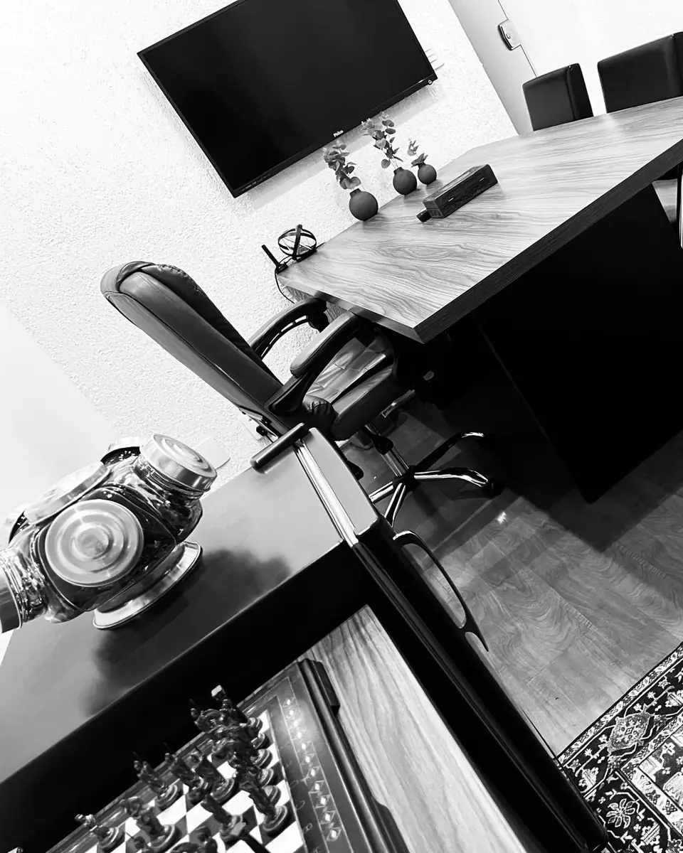 Sala de reunião do escritório da Dra. Cinthya Andrade, em preto e branco: mesa de madeira, cadeira de couro, televisão na parede e um tabuleiro de xadrez